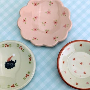 May include: Three decorative ceramic plates. One is pink with a scalloped edge and floral pattern. Another is light green with a black cat wearing a Santa hat. The third is light green with a red heart pattern.