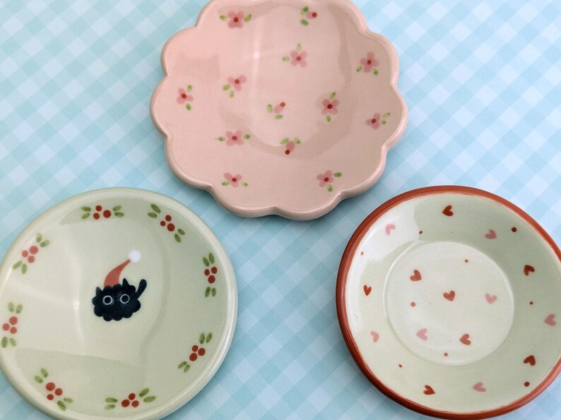 May include: Three decorative ceramic plates. One is pink with a scalloped edge and floral pattern. Another is light green with a black cat wearing a Santa hat. The third is light green with a red heart pattern.
