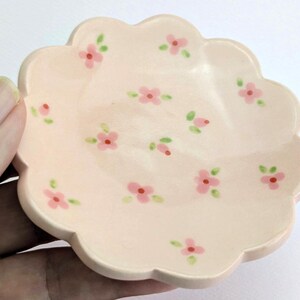 May include: A small, scalloped-edge, ceramic dish in a soft peach color. The dish is decorated with hand-painted pink flowers and green leaves. The dish is held in a hand, and is likely used for jewelry or small items.