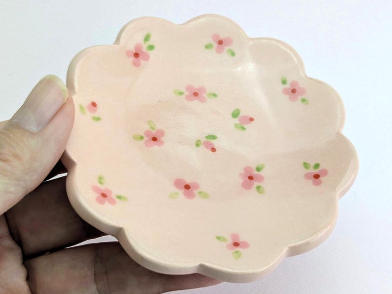 May include: A small, scalloped-edge, ceramic dish in a soft peach color. The dish is decorated with hand-painted pink flowers and green leaves. The dish is held in a hand, and is likely used for jewelry or small items.