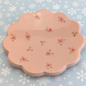 May include: A light pink, scalloped-edge ceramic dish decorated with small pink flowers and green leaves. The dish is on a light blue surface with white snowflake patterns.