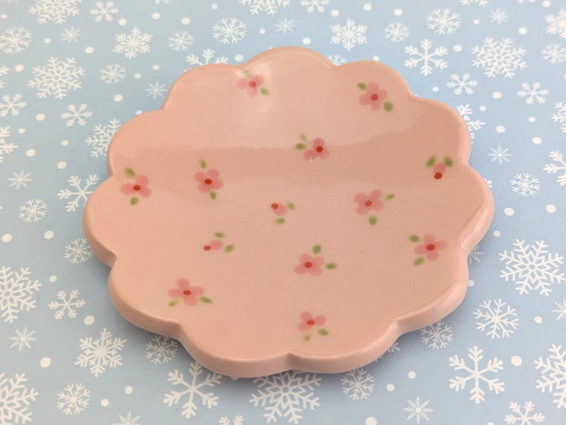 May include: A light pink, scalloped-edge ceramic dish decorated with small pink flowers and green leaves. The dish is on a light blue surface with white snowflake patterns.