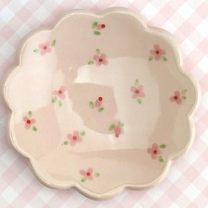 May include: A light pink, scalloped-edge ceramic dish decorated with small pink flowers and green leaves. The dish has a shallow bowl shape and is likely used for serving small portions of food or as a decorative item.