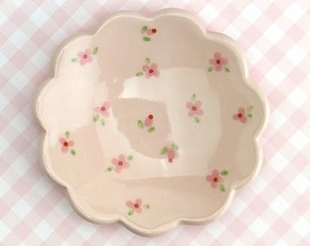 May include: A pale pink ceramic dish with a scalloped edge, adorned with hand-painted pink flowers and green leaves. The dish is designed with a shallow bowl and is suitable for serving small portions or as a decorative piece.