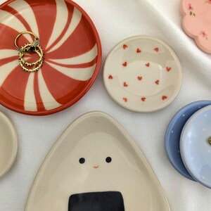 May include: A collection of decorative ceramic dishes in various shapes and designs. Includes a red and white swirled dish holding gold rings, a small heart-patterned dish, and a light blue dish with a floral accent. Other dishes feature floral and onigiri designs.