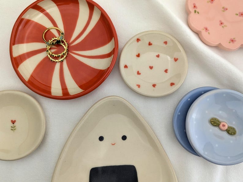 May include: A collection of decorative ceramic dishes in various shapes and designs. Includes a red and white swirled dish holding gold rings, a small heart-patterned dish, and a light blue dish with a floral accent. Other dishes feature floral and onigiri designs.