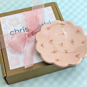 May include: A pink, scalloped-edge ceramic dish decorated with small pink flowers and green leaves. The dish sits next to a brown box tied with a pink ribbon and a tag with the name "chris".