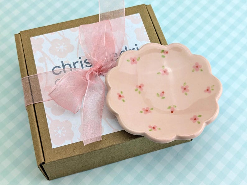May include: A pink, scalloped-edge ceramic dish decorated with small pink flowers and green leaves. The dish sits next to a brown box tied with a pink ribbon and a tag with the name "chris".