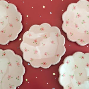 May include: Five pink ceramic plates with scalloped edges are arranged on a red surface. Each plate is decorated with small pink floral patterns. The plates are a light pink color, and the flowers have green leaves.