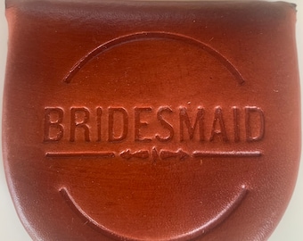 Könnte beinhalten: Eine braune Ledermünzbörse mit dem Wort "BRIDESMAID" auf der Vorderseite geprägt.