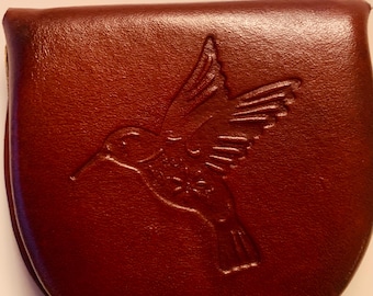 Könnte beinhalten: Eine dunkelbraune Münzbörse aus Leder mit einem eingeprägten Kolibri-Motiv auf der Vorderseite. Das Täschchen hat eine abgerundete, halbkreisförmige Form mit glatten, polierten Kanten und einer sichtbaren Falte oben am Verschluss.