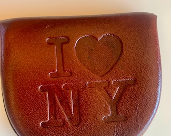 Könnte beinhalten: Eine braune Leder-Münzbörse mit dem geprägten Schriftzug "I ❤️ NY". Die Börse hat einen gebogenen Boden und einen Klappenverschluss. Die Buchstaben und das Herz sind leicht erhaben und erzeugen einen strukturierten Effekt.