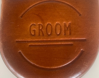 Könnte beinhalten: Eine braune Ledertasche mit dem Wort "GROOM" auf der Vorderseite geprägt. Die Tasche hat eine gebogene Form und einen genähten Rand. Der Text befindet sich in einem gebogenen Design.
