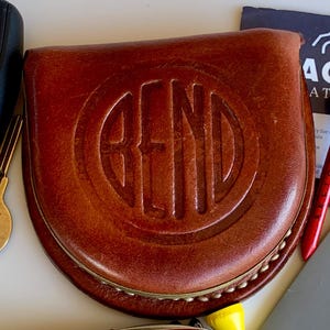 May include: A brown leather golf putter cover with a rounded shape and a debossed circular logo featuring the word BEND in the center. The cover has visible light-colored stitching along the bottom edge and a smooth, polished finish on the leather surface.