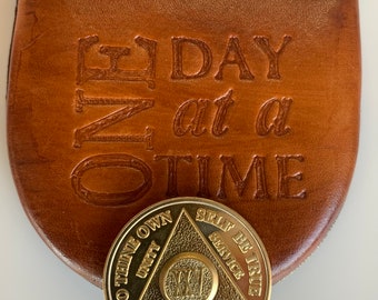 Könnte beinhalten: Eine goldene Münze mit einem Dreiecksdesign und den Worten "To Thine Own", "Unity", "Recovery", "Self Be True" und "Service", die um das Dreieck herum eingraviert sind. Die Münze liegt auf einer braunen Ledertasche mit den geprägten Worten "One Day at a Time".