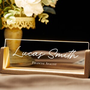 Puede incluir: Un letrero de escritorio de madera con un panel de acrílico transparente iluminado desde atrás. El panel muestra el nombre "Lucas Smith" en escritura cursiva blanca y el título "FINANCIAL ANALYST" en texto blanco más pequeño.