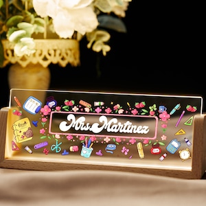 May include: A wooden desk nameplate with a clear acrylic insert featuring a pink and white floral design with a pink banner that reads "Mrs. Martinez". The acrylic insert is illuminated with warm white LED lights. The design includes various school-themed icons such as pencils, a ruler, a calculator, a book, and a cup of coffee.