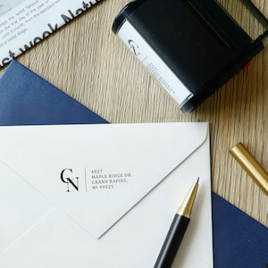 Op de afbeelding: Een witte envelop met een zwarte, gepersonaliseerde stempel met het retouradres, de initialen CN en de tekst 4827 MAPLE RIDGE DR. GRAND RAPIDS, MI 49525. De envelop ligt op een donkerblauw vel karton naast een stempel en een gouden pen.