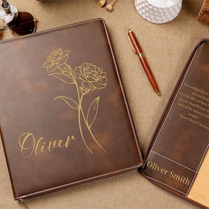 Könnte beinhalten: Ein braunes Lederjournal mit floralem Design und dem Namen "Oliver" in goldener Schrift. Das Journal ist geöffnet und zeigt ein beiges Innere und ein Zitat. Ein Stift und eine Sonnenbrille sind ebenfalls sichtbar.