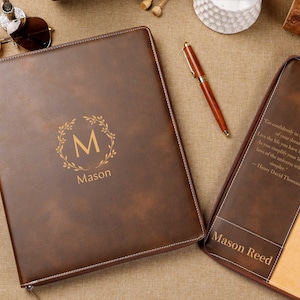 以下が含まれることがあります： 「Mason」の名前と「M」のイニシャルがリースデザインで施された茶色の革製ポートフォリオ。ポートフォリオは開いており、ベージュの内側が見えます。ペンとサングラスも表示されています。