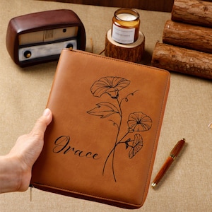 May include: A brown leather journal with a floral design and the name "Grace" in black script. The journal has a zipper closure and is held by a person. A pen and other items are in the background.