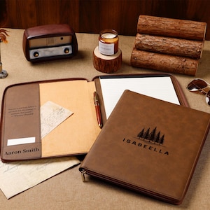 May include: A brown leather portfolio with the name "Isabella" and a tree graphic, open to reveal a notepad and pen. A vintage radio, candle, sunglasses, and compass are also present.