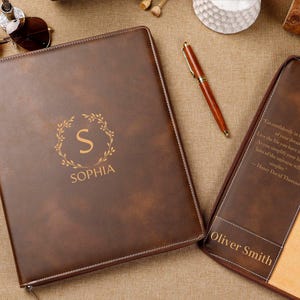 Puede incluir: Portafolio de cuero marrón con el nombre "SOPHIA" y un diseño de corona dorada. Un portafolio abierto a juego muestra el nombre "Oliver Smith" y una cita. También se ven un bolígrafo y gafas de sol.