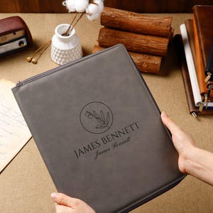 May include: A gray leather portfolio with a zipper closure. The portfolio features the name "JAMES BENNETT" and a circular olive branch design. The portfolio is held open, showcasing the design. The background includes a vase with cotton stems and other leather-bound items.
