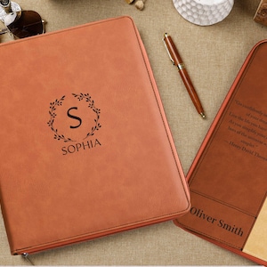 Peut inclure: Un portfolio en cuir marron clair personnalisé avec le nom "SOPHIA" et une couronne de feuilles avec la lettre "S". Un stylo assorti et un second portfolio avec le nom "Oliver Smith" sont également visibles.