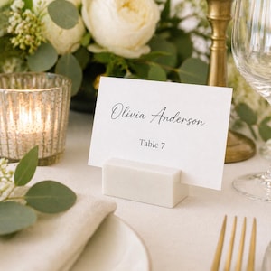 May include: A white place card with the name "Olivia Anderson" and "Table 7" printed in black script. The card is held in a rectangular, off-white holder. The setting includes a gold fork, a candle, and a floral arrangement.