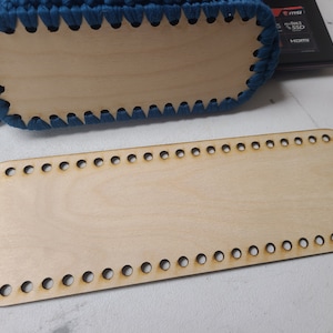 Puede incluir: Una base de madera de color marrón claro con forma rectangular redondeada y agujeros en los bordes. La base está diseñada para proyectos de ganchillo o tejido.