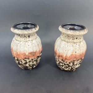 May include: Two ceramic vases with a textured, layered design. The vases have a dark brown base, a band of orange-red, and a speckled beige top. The rims are dark brown. The vases are displayed on a dark surface.