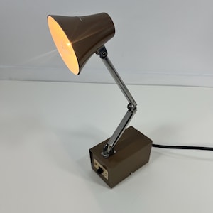 May include: A brown desk lamp with a pivoting arm and a rectangular base. The lamp shade is angled, and the light is on. The lamp has a silver-colored, adjustable arm and a switch on the base.