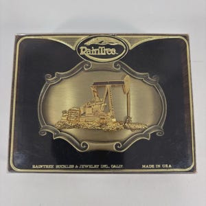 May include: A rectangular belt buckle with a gold-colored oil derrick design on a dark brown background. The buckle has the text "Raintrees" and "Raintree Buckles & Jewelry Inc., Calif." and "Made in USA".