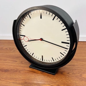 May include: A black, round clock with a cream-colored face and black hands. The clock has a black frame and a black base. The clock's minute and hour markers are black. The second hand is red.