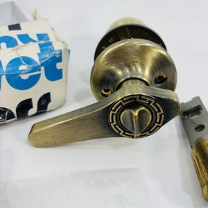 May include: An antique brass-finished door handle with a lever design and a circular base featuring a Greek key pattern. The handle includes a central heart-shaped thumb turn and is accompanied by a metal latch mechanism and a partial cardboard box.