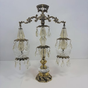 May include: An ornate, vintage-style lamp with a brass finish and crystal accents. The lamp features three light fixtures with glass shades and hanging crystals. The base has a marble accent and decorative detailing, creating an elegant aesthetic.