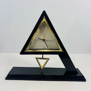 May include: A triangular black and gold mantel clock featuring a cream-colored face with a grid pattern, a Roman numeral XII at the top, and black hands. A small gold triangular pendulum hangs below the main frame on a black rectangular base.