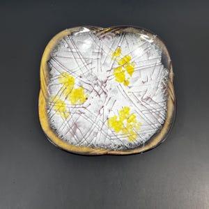 May include: Square decorative dish with a white interior featuring yellow floral accents and thin, dark lines. The rim is a mix of gold and brown, creating a vintage aesthetic. The dish is set against a dark background.
