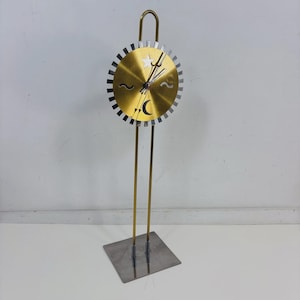 Peut inclure: Une horloge décorative de couleur or avec un design unique. Le cadran de l'horloge présente une étoile, un croissant de lune et des symboles de vagues. L'horloge est soutenue par deux fines tiges dorées et une base carrée argentée. Le haut de l'horloge est incurvé.