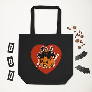 Può includere: Borsa tote nera con un design a tema Halloween. Il design presenta una zucca con una creatura simile a un pipistrello all'interno di un cuore rosso. La parola "BOO" è scritta in singole lettere nere a sinistra.