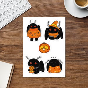 Può includere: Un foglio di adesivi con falene dei cartoni animati in costume di Halloween. Le falene sono nere con occhi rossi e indossano maglioni, cappelli e sciarpe arancioni. Una falena tiene in mano una zucca intagliata. È incluso anche un adesivo a forma di zucca.