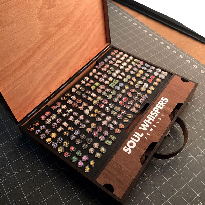 May include: A wooden jewelry box with a hinged lid, filled with numerous colorful enamel pins. The box has a dark brown exterior and a lighter wood interior. The text "SOUL WHISPERS JEWELRY" is visible on the side.