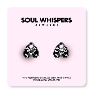 May include: Black Ouija board-shaped earrings with the words "HELLO" and "GOODBYE" in white. The earrings also feature the words "YES" and "NO". The earrings are on a pink background with the text "SOUL WHISPERS JEWELRY".