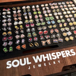 May include: A collection of colorful, cartoon-themed charms, including characters like Spider-Man, Batman, and Kermit the Frog, arranged on a dark wooden surface. The text "SOUL WHISPERS JEWELRY" is visible in white. These charms are likely for jewelry making.