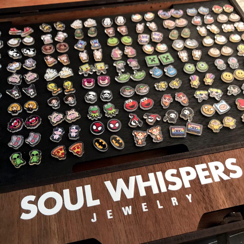 May include: A collection of colorful, cartoon-themed charms, including characters like Spider-Man, Batman, and Kermit the Frog, arranged on a dark wooden surface. The text "SOUL WHISPERS JEWELRY" is visible in white. These charms are likely for jewelry making.
