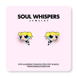 May include: A pair of cartoon character earrings featuring a blonde-haired girl with blue eyes, black and white outfit, and a smiling expression. The earrings are on a pink background with the text "SOUL WHISPERS JEWELRY". The earrings are made with hypo-allergenic stainless steel posts and backs.
