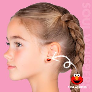 May include: A young person with blonde hair wearing a pair of red Elmo stud earrings. The earrings feature the character's face with black eyes and a wide smile. The background is a solid pink color.