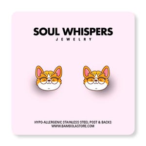 May include: A pair of corgi-shaped stud earrings with orange and white details, wearing round glasses. The earrings are displayed on a pink card with the text "SOUL WHISPERS JEWELRY" and "HYPO-ALLERGENIC STAINLESS STEEL POST & BACKS".