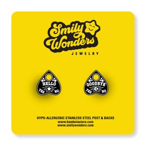 May include: A pair of black and white planchette-shaped stud earrings on a bright yellow display card. The left earring features the text HELLO, YES, and NO with a star. The right earring features GOODBYE, YES, and NO with a moon and star.